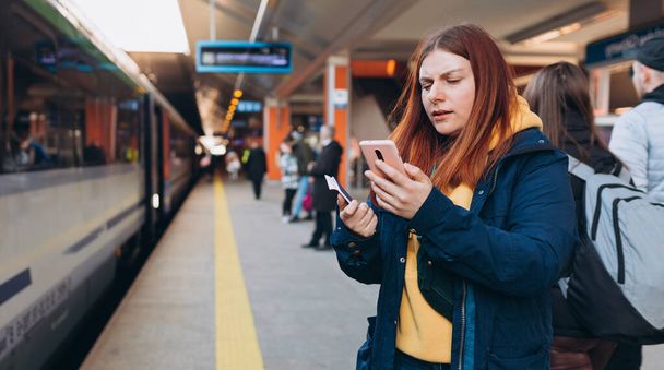 Junge rothaarige Frau wartet mit Rucksack auf Bahnsteig und Smartphone. Eisenbahnverkehrskonzept, Reisende. Invasion in der Ukraine. - Foto, Bild