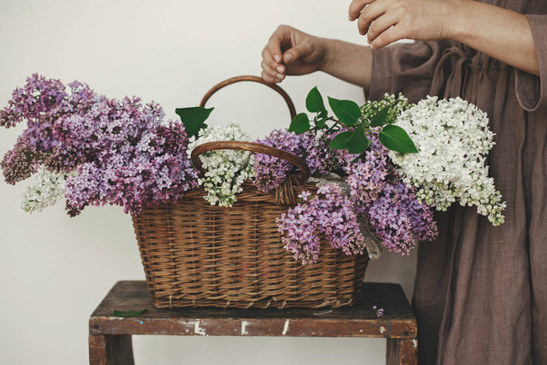 Stílusos nő gyönyörű lila virágok fonott kosárban rusztikus szobában. Nő vászonban ruha elrendezése orgona virágok a vidéki szobában, vágott kilátás. Eredeti tavaszi pillanatok - Fotó, kép