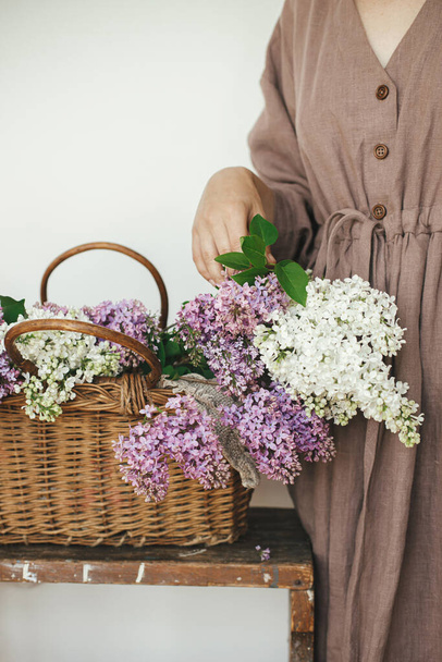 Mulher elegante com belas flores lilás na cesta de vime no quarto rústico. Feminino em vestido de linho organizando flores lilás no quarto rural, vista cortada. Momentos autênticos da primavera - Foto, Imagem