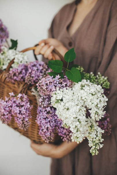 Stylish woman holding wicker basket with beautiful lilac flowers in rustic room. Female in linen dress arranging lilac flowers, cropped view. Authentic moody moment. Rustic wedding - 写真・画像