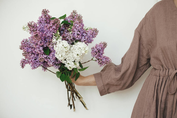 Stylish woman holding beautiful lilac flowers against white wall. Female in linen dress with lilac flowers, cropped view. Authentic moody moment. Rustic wedding - Фото, изображение