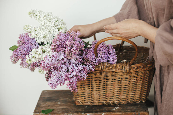 Stylish woman with beautiful lilac flowers in wicker basket in rustic room. Female in linen dress arranging lilac flowers in rural room, cropped view. Authentic spring moments - Фото, изображение