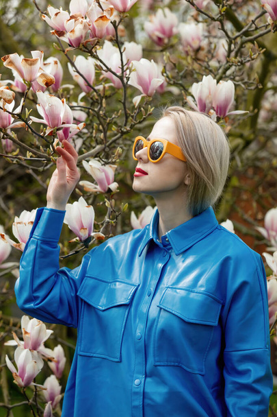 Beautiful blond woman in sunglasses and blue shirt next to Magnolia tree in spring time - Fotografie, Obrázek