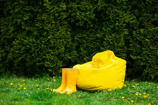 Soft Bag Stuhl und gelbe Stiefel auf dem Rasen mit Löwenzahn auf einem Garten  - Foto, Bild