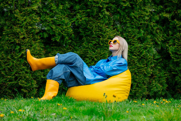 Stylish ukrainian woman in blue shirt and yellow boot sits on soft chair in spring garden  - Foto, Imagen