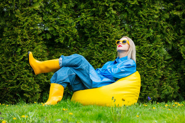 Stylish ukrainian woman in blue shirt and yellow boot sits on soft chair in spring garden  - Valokuva, kuva