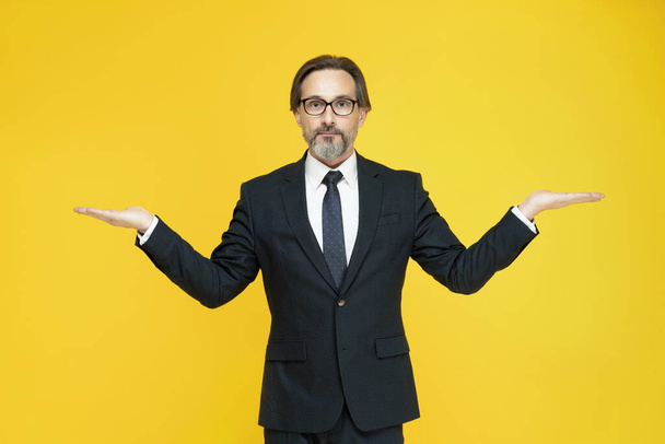 Weighing or balancing middle aged business man with hands stretched sideways looking at camera isolated on yellow background. Handsome mature businessman in black suit. Business concept. Copy space - Photo, image
