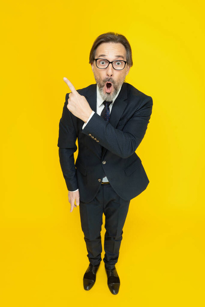 Funny excited middle aged business man showing sideways looking at camera isolated on yellow background. Handsome mature businessman in black suit. Business concept. Copy space - Фото, изображение