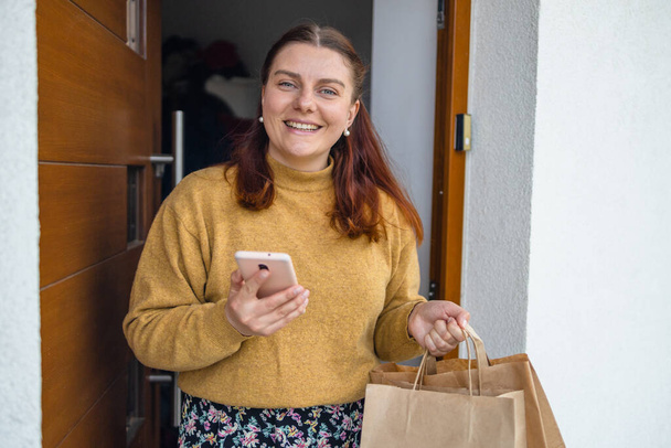 Home delivery. Woman with smartphone opening house entrance door while meeting courier with online order, postwoman delivering package to customer. - Photo, image