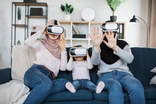 Three generations of islamic women in hijab in VR glasses sitting on sofa at home. - Foto, Imagem