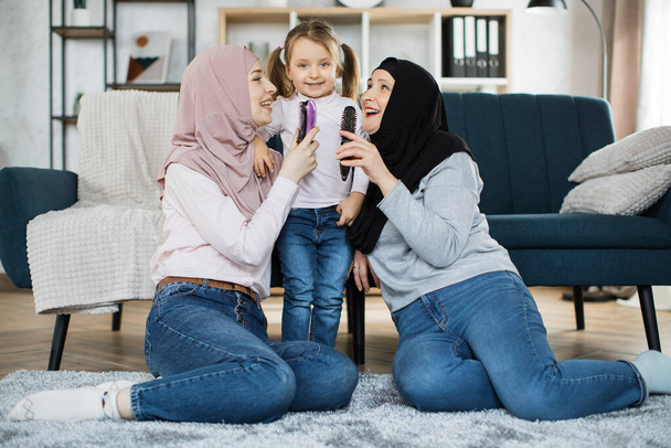 Three generations of Muslim women sitting on their knees on floor near the sofa in the living room - Фото, изображение