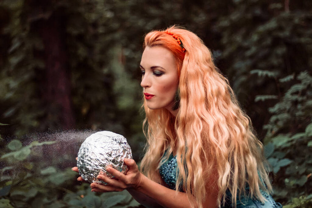 Una joven hermosa mujer con el pelo rojo sopla polvo de una bola mágica - Foto, imagen