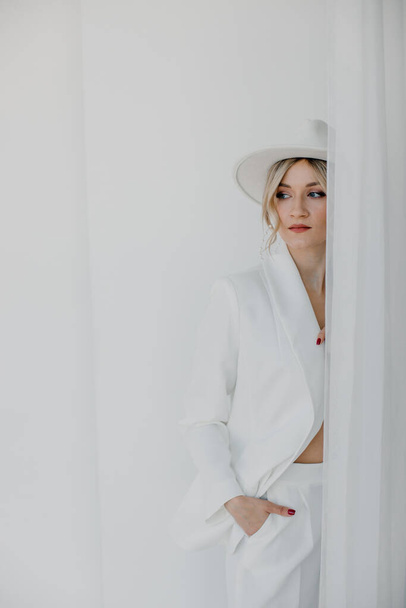 Confident young blond woman smiling, looking at camera isolated on white background. Studio portrait of successful friendly female in white suit and hat, posing over white wall. - Photo, image