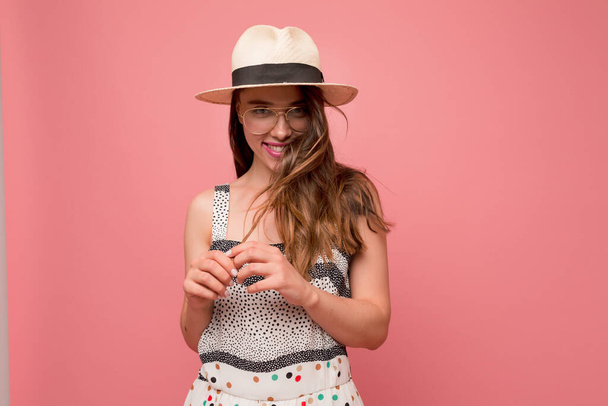 Stylish trendy girl with brown wavy hair wearing light hat and glasses and summer dress is smiling at camera and having fun over pink background. High quality photo - Foto, imagen