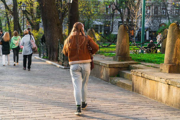Молодая свободная девушка с длинными волосами и косичкой идет по весенней улице в свете вечернего солнца с белыми наушниками на голове позади группы взрослых женщин с сумками для покупок - Фото, изображение