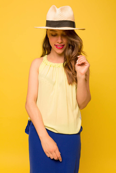 Elegant lovely girl with wavy brown hair wearing hat and yellow with blue dress is looking down and smiling with shy smile. High quality photo - Foto, afbeelding