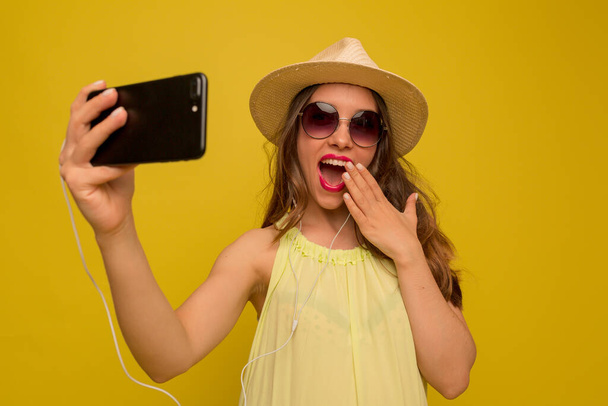Garota encantadora excitada com cabelos longos ondulados em óculos de sol e chapéu está cobrindo os lábios com a mão e fazendo selfie no smartphone. Foto de alta qualidade - Foto, Imagem