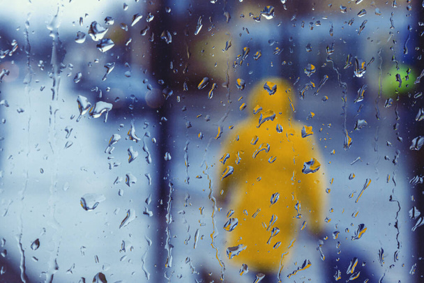 Sarı bir yağmurluk giyen yayalar yağmurda yürüyor, ön planda yağmur damlaları var. - Fotoğraf, Görsel
