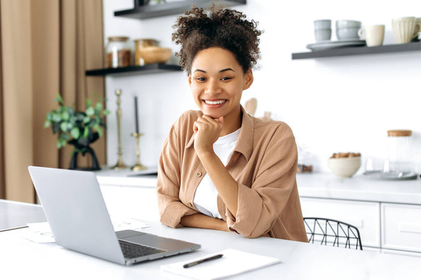 Portrait of a pretty african american brunette woman. Successful female freelancer, student, designer, wearing casual shirt, sit at home in the kitchen with laptop, looks at camera, smiling friendly - Foto, Imagen