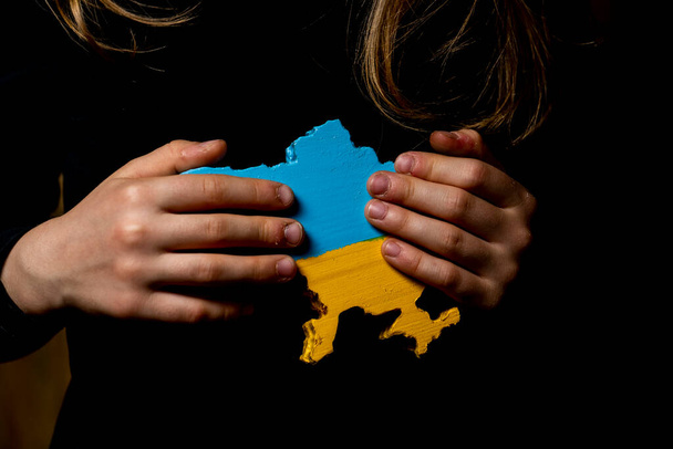 a map of Ukraine carved from wood and painted in the colors of the Ukrainian flag in the hands of a girl on a dark background - Foto, Bild