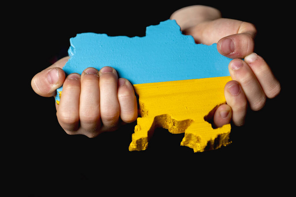 mapa Ukrajiny vytesaná ze dřeva a malovaná v barvách ukrajinské vlajky v rukou dívky na tmavém pozadí - Fotografie, Obrázek