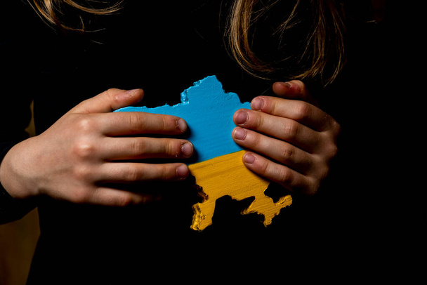 a map of Ukraine carved from wood and painted in the colors of the Ukrainian flag in the hands of a girl on a dark background - Фото, зображення