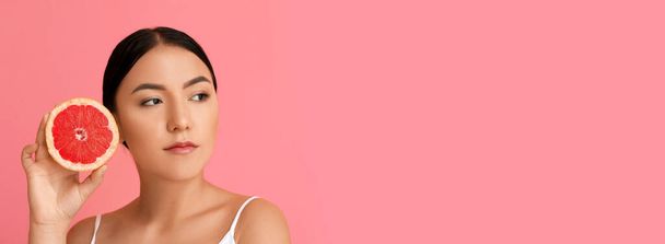 Beautiful young Asian woman holding grapefruit on pink background with space for text - Фото, изображение