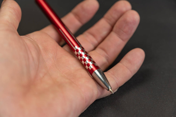 Um close-up da mão de um homem segurando uma caneta vermelha contra um fundo preto. Caneta esferográfica de metal deitada na palma da mão de um homem maduro. - Foto, Imagem