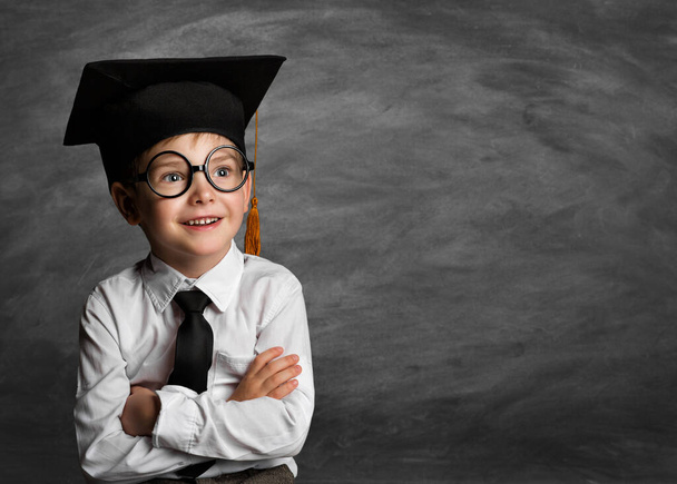 Happy Child in Glasses and Graduation Hat thinking over Blackboard. Cheerful smiling Preschool Boy in Eyeglasses over Chalkboard. Clever Kids Elementary Education - 写真・画像