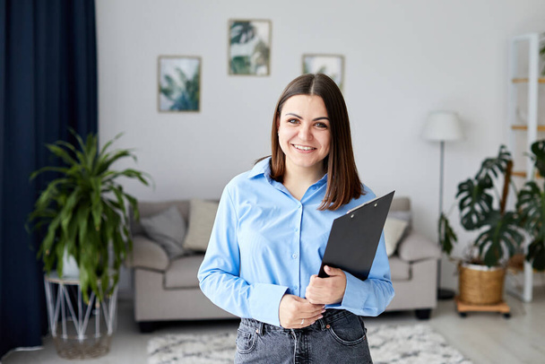 A young pretty girl is standing in a cozy living room or classroom and holding a black folder. Woman ready to work and smiling happily - Valokuva, kuva