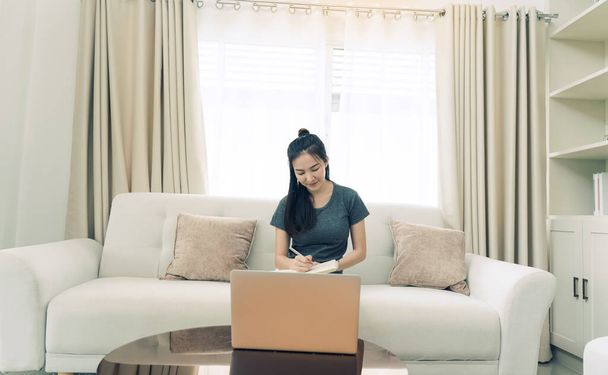 Mujer asiática se sienta en el sofá mientras toma notas de la pantalla de su computadora mientras trabaja o estudia en línea en casa. - Foto, Imagen