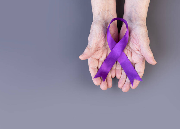 Hands of elderly woman hold purple ribbon on gray background. Alzheimer's awareness concept. Copy space. - Foto, Imagem