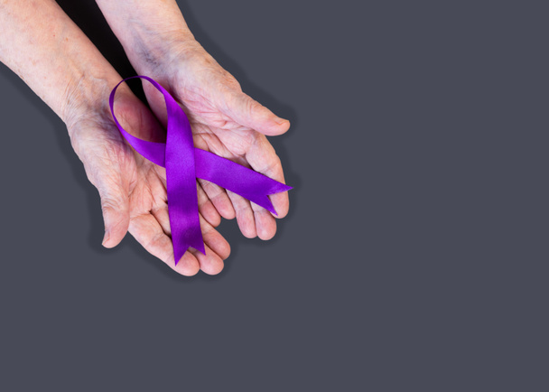 Hands of elderly woman hold purple ribbon on gray background. Alzheimer's awareness concept. Copy space. - Foto, imagen
