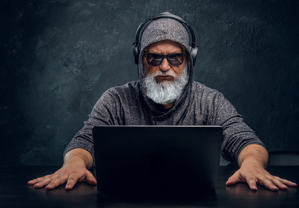 Tricky ηλικιωμένος άνδρας με κουκούλα και ακουστικά χρησιμοποιώντας φορητό υπολογιστή - Φωτογραφία, εικόνα