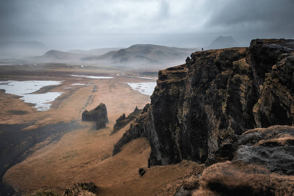 Musta hiekkaranta, sammalta peitetty kallioita ja vuoria Dyrholaey, Etelä-Islanti, syksyllä. Tunnistamaton valokuvaaja on siluetoituna kalliolle. - Valokuva, kuva