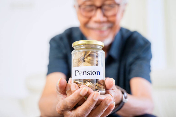 Happy old man showing a jar full of coins with pension word while sitting on the couch - Фото, изображение