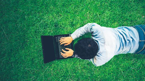 Top down view of young man using a laptop while lying on the green grass at the park - Foto, Bild