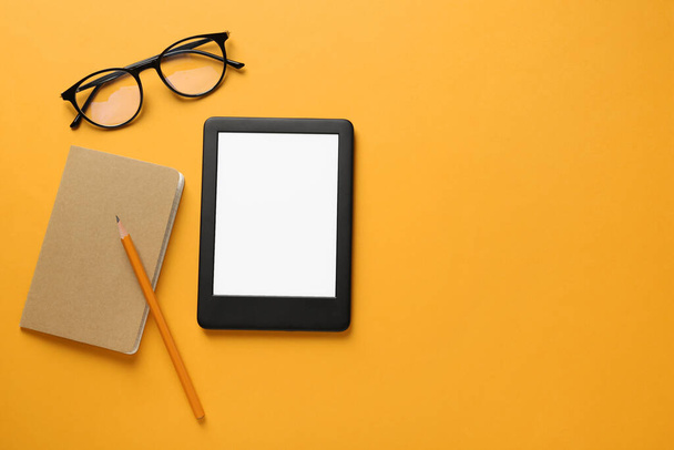 Modern e-book reader, glasses, notebook and pencil on orange background, flat lay. Space for text - Φωτογραφία, εικόνα