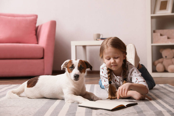 Cute little girl with her dog reading book on carpet at home. Childhood pet - Фото, зображення