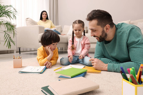 Father playing with his children while mother reading book on sofa in living room - Photo, Image
