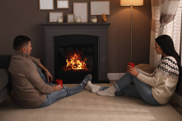 Lovely couple with cups of hot drink spending time together near fireplace at home - Фото, изображение