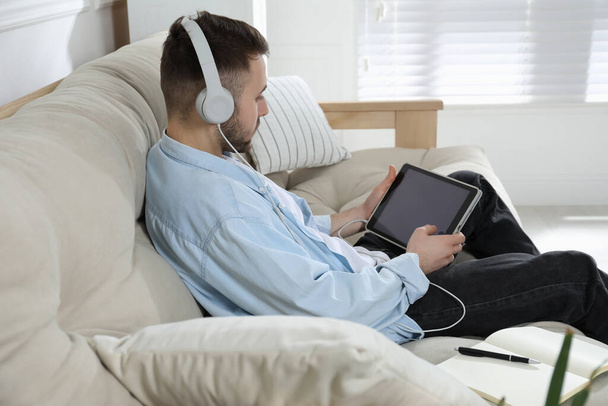 Young man with headphones using modern tablet for studying on sofa at home. Distance learning - Foto, Imagem
