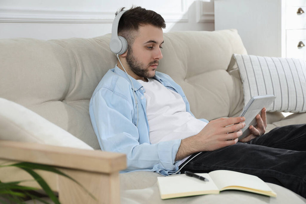 Young man with headphones using modern tablet for studying on sofa at home. Distance learning - Foto, immagini