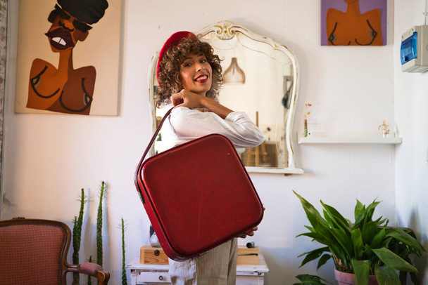 Молодая, красивая, богемная латиноамериканка с красным беретом на голове и красным винтажным чемоданом, счастливо смотрящая в камеру для фотографии. Концепция предприимчивой женщины. - Фото, изображение