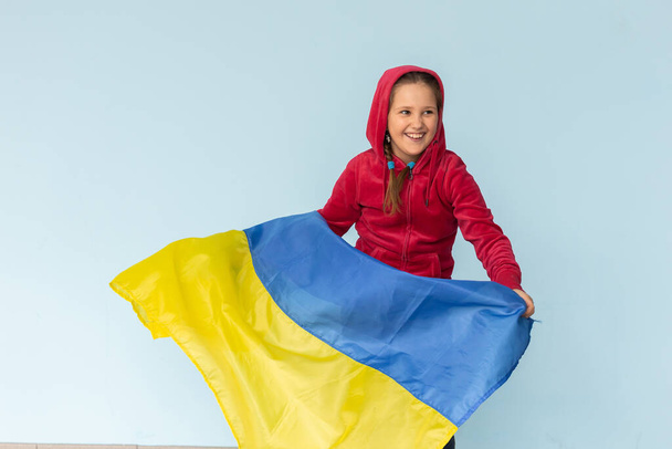 Flagge der Ukraine in den Händen eines glücklichen ukrainischen Mädchens. Kind lächelt in blau-gelber Fahne der Ukraine, die am blauen Himmel flattert. - Foto, Bild