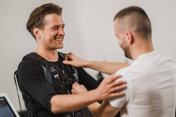 Osobní trenér pomáhá muži středního věku s dressingem pro EMS cvičení v tělocvičně. - Fotografie, Obrázek