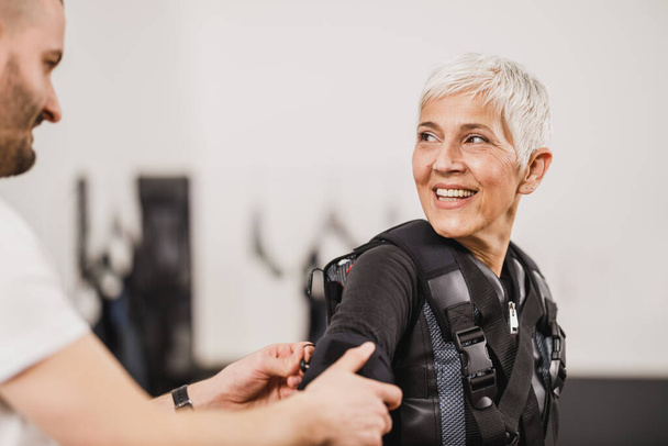 Osobní trenér pomáhá žena středního věku s dressingu pro EMS cvičení v tělocvičně. - Fotografie, Obrázek