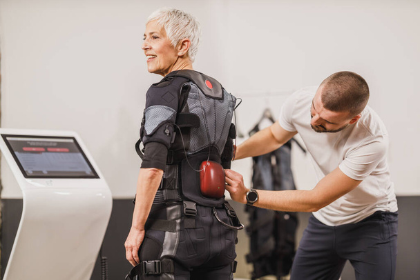 Personal Trainer hilft einer Frau mittleren Alters im Anzug beim EMS-Training im Fitnessstudio. - Foto, Bild