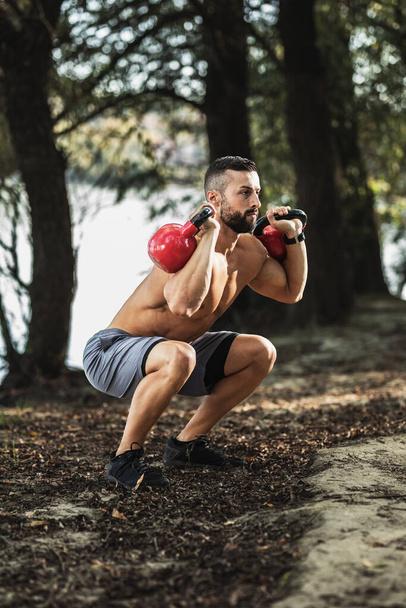 Muskulöser junger Mann mit nacktem Oberkörper macht Kniebeuge-Übungen mit der Kettlebell in der Natur, in der Nähe des Flusses. - Foto, Bild