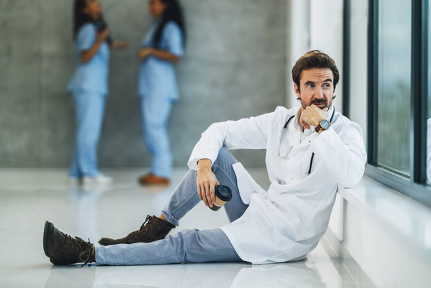 Un médecin inquiet assis sur le sol et cher regardant par la fenêtre tout en faisant une pause café rapide dans un couloir vide de l'hôpital. - Photo, image